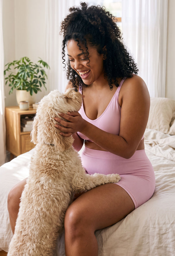 Smiling woman sitting on a bed cuddling a fluffy cream dog, wearing Lilac Snow cotton rich pyjama sleep shorts with matching lilac lounge bralette