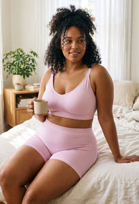 Woman sitting on the edge of a bed holding a mug, wearing Lilac Snow cotton rich pyjama sleep shorts with matching lilac lounge bralette